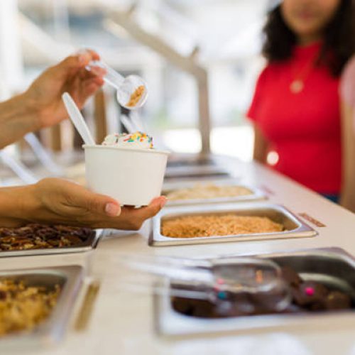 Close up of woman hands holding delicious ice cream or frozen yogurt at the toppings bar and selling to teen girls customers